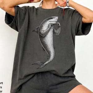 May include: A charcoal gray short-sleeve t-shirt featuring a black and white graphic illustration of a shark standing upright and playing a harmonica. The shirt has a relaxed fit and a crew neckline. The text Comfort Colors Pepper is visible.