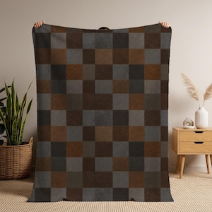 May include: A large, rectangular blanket with a patchwork design. The blanket features a repeating pattern of squares in shades of brown, gray, and black. The blanket is held up by unseen hands, showcasing its size and design. The blanket is made of a soft material.