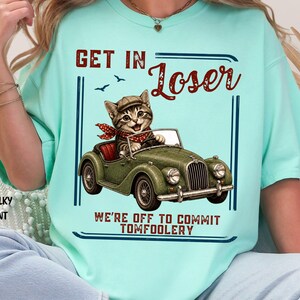 May include: A chalky mint t-shirt featuring a graphic of a tabby cat wearing a newsboy cap and red polka dot scarf, driving a vintage green convertible car. The text reads "GET IN LOSER" and "WE'RE OFF TO COMMIT TOMFOOLERY" in a dark red font.