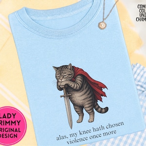 May include: A light blue t-shirt featuring a graphic of a tabby cat wearing a red cape and holding a sword. The text below the cat reads, "alas, my knee hath chosen violence once more." The shirt includes a small necklace and text labels.
