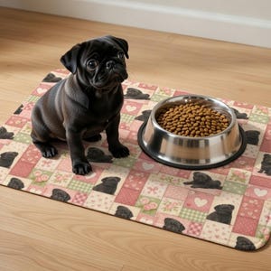 May include: A black pug sits on a pink and green patchwork pet feeding mat. The mat features a repeating pattern of pug illustrations, hearts, and floral designs. A stainless steel bowl filled with dog food sits on the mat.