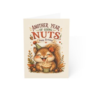 Puede incluir: Una tarjeta de cumpleaños color crema con un dibujo de una ardilla sonriente que lleva un gorro de fiesta verde con lunares. La ardilla sostiene un cupcake con una vela. El texto dice: ANOTHER YEAR OF GOING NUTS Happy Birthday! Rodeada de bellotas.