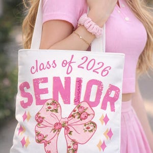 May include: White tote bag with pink text reading "class of 2026 SENIOR Savannah" and a floral bow graphic. The bag is held by a person wearing a pink outfit. The bag has a white strap.