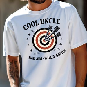 May include: White t-shirt with the text "COOL UNCLE" above a bullseye target design with three darts. Below the target, the text reads "BAD AIM • WORSE ADVICE". The shirt is a comfort colors style.
