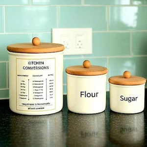 May include: Three off-white kitchen canisters with wooden lids and knobs. The largest canister has "Kitchen Conversions" text. The other two are labeled "Flour" and "Sugar". The canisters sit on a black surface against a teal tile background.