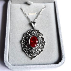 May include: A silver-toned necklace featuring an oval-shaped red gemstone pendant set within an intricate, openwork metal frame. The pendant hangs from a delicate silver-toned chain, showcasing detailed filigree patterns throughout the design.