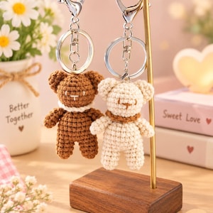 May include: Two crochet bear keychains hanging from a gold-toned metal stand. One bear is dark brown with a white collar, and the other is cream-colored with a brown collar. Both feature silver-toned metal lobster clasps and circular key rings.