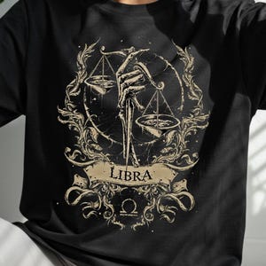 Op de afbeelding: Zwart T-shirt met een Weegschaal sterrenbeeld ontwerp. De afbeelding toont een skelet hand die een weegschaal vasthoudt, omringd door sierlijke krullen. Het woord "LIBRA" staat op een banner.