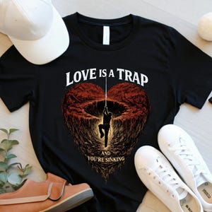 May include: Black t-shirt with the text "LOVE IS A TRAP and YOU'RE SINKING" and a graphic of a person hanging from a rope in a heart-shaped pit. The image also includes a white baseball cap, sunglasses, white sneakers, and a pair of brown shoes.