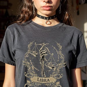 Op de afbeelding: Donkergrijs t-shirt met een Weegschaal sterrenbeeld ontwerp. De afbeelding toont een skeletachtige hand die een weegschaal vasthoudt, omringd door sierlijke bloemmotieven. Het woord "LIBRA" staat op een banner onderaan het ontwerp.