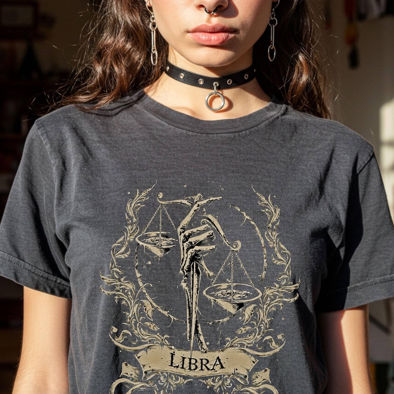 Op de afbeelding: Donkergrijs t-shirt met een Weegschaal sterrenbeeld ontwerp. De afbeelding toont een skeletachtige hand die een weegschaal vasthoudt, omringd door sierlijke bloemmotieven. Het woord "LIBRA" staat op een banner onderaan het ontwerp.