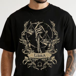 Op de afbeelding: Zwart T-shirt met een Weegschaal sterrenbeeld ontwerp. De afbeelding toont een skelet hand die een weegschaal vasthoudt, omringd door sierlijke bloemmotieven en het woord "LIBRA" op een banner. Het ontwerp is in beige en bruine tinten.