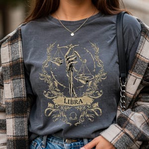 Op de afbeelding: Donkergrijs t-shirt met een goudkleurig Weegschaal sterrenbeeld ontwerp. Het ontwerp toont een skelet hand die een weegschaal vasthoudt, omringd door sierlijke bloemelementen en het woord "LIBRA".