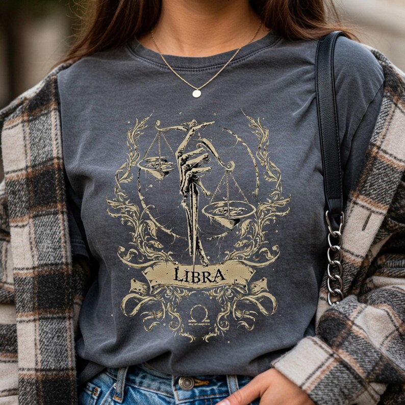Op de afbeelding: Donkergrijs t-shirt met een goudkleurig Weegschaal sterrenbeeld ontwerp. Het ontwerp toont een skelet hand die een weegschaal vasthoudt, omringd door sierlijke bloemelementen en het woord "LIBRA".