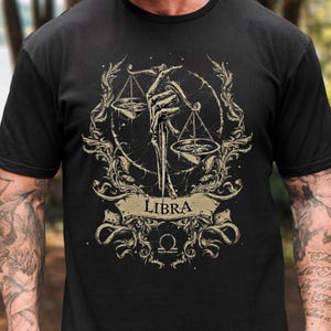 Op de afbeelding: Zwart T-shirt met een Weegschaal sterrenbeeld ontwerp. Het ontwerp toont een skeletachtige hand die een weegschaal vasthoudt, omringd door sierlijke krullen en het woord "Libra" op een spandoek. De weegschaal en het spandoek zijn lichtbruin.