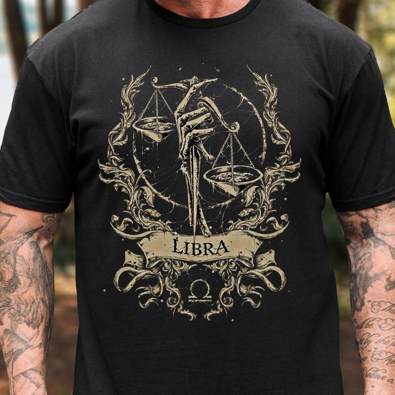 Op de afbeelding: Zwart T-shirt met een Weegschaal sterrenbeeld ontwerp. Het ontwerp toont een skeletachtige hand die een weegschaal vasthoudt, omringd door sierlijke krullen en het woord "Libra" op een spandoek. De weegschaal en het spandoek zijn lichtbruin.