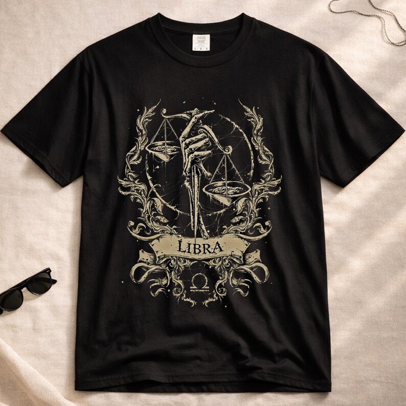 Op de afbeelding: Zwart T-shirt met een Weegschaal sterrenbeeld ontwerp. De afbeelding toont een skelet hand die een weegschaal vasthoudt, omringd door sierlijke krullen. Het woord "LIBRA" staat op een banner onder de weegschaal, met het Weegschaal symbool eronder.