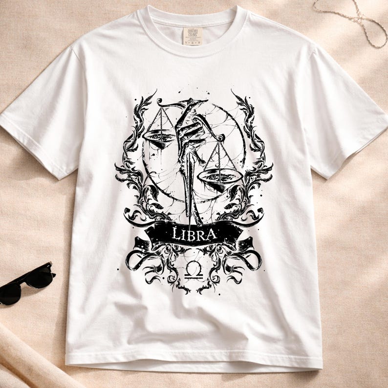 Op de afbeelding: Wit T-shirt met een zwart Weegschaal sterrenbeeld ontwerp. De afbeelding toont een weegschaal, decoratieve elementen en het woord "Libra" op een banner. Het shirt heeft korte mouwen en een ronde hals.