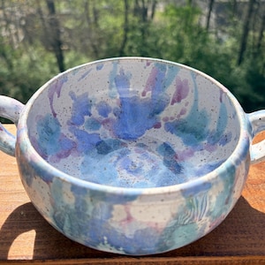 Può includere: Una zuppiera in ceramica con due manici, con interno bianco maculato e un design ad acquerello nei toni del blu, verde acqua e viola. La zuppiera è appoggiata su una superficie di legno, con sfondo naturale.