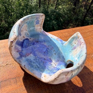 Może przedstawiać: Ręcznie robiona ceramiczna miska o unikalnym kształcie półksiężyca. Misa ma mieszankę niebieskich, fioletowych i białych glazur, tworząc efekt marmuru. Obrzeże ma teksturowany, nakrapiany wygląd, dodający uroku rzemieślniczego.