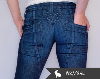 May include: Dark blue denim jeans with a decorative design on the back pockets and a unique emblem above the back pockets. The jeans have a straight leg fit. The tag shows W27/35L.