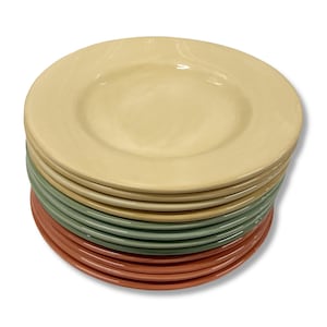 Pode incluir: Uma pilha de pratos de cerâmica em várias cores, incluindo creme, verde e terracota. Os pratos têm um design simples e redondo com uma borda ligeiramente elevada. Os pratos estão empilhados de forma organizada.