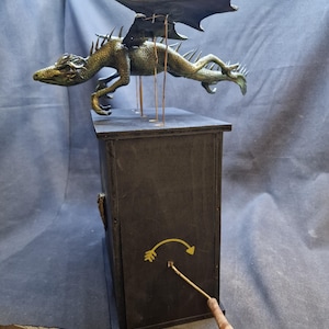 May include: A bronze dragon sculpture with outstretched wings, mounted on a black box. The dragon features detailed scales and spikes. A winding key is inserted into the box, with a yellow arrow indicating its function.