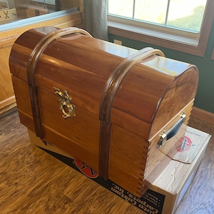 May include: A wooden trunk with a curved top and brass hardware. The trunk is a warm brown color with darker brown accents and a black handle. The trunk is sitting on a cardboard box.