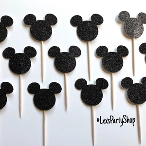 Pode incluir: Doze toppers para cupcake com cabe&ccedil;a de Mickey Mouse em glitter preto em palitos de madeira. Os toppers est&atilde;o dispostos em uma grade sobre um fundo branco.