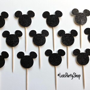 Pode incluir: Doze toppers para cupcakes com a forma de cabe&ccedil;a do Mickey Mouse em glitter preto em palitos de madeira. Os toppers est&atilde;o dispostos em c&iacute;rculo sobre um fundo branco. @LexsPartyShop est&aacute; escrito no canto inferior direito.