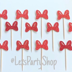 May include: Twelve red glitter bow cupcake toppers on wooden sticks. The bows are arranged in a grid pattern on a white background. The text '@LexsPartyShop' is visible in the bottom center of the image.