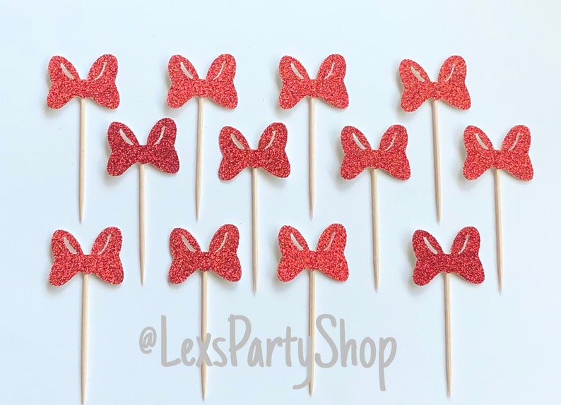 May include: Twelve red glitter bow cupcake toppers on wooden sticks. The bows are arranged in a grid pattern on a white background. The text '@LexsPartyShop' is visible in the bottom center of the image.