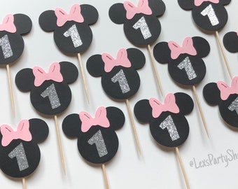 May include: Twelve black and pink Minnie Mouse themed cupcake toppers with silver glitter number ones. The toppers are on wooden skewers.