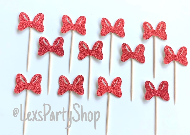May include: Twelve red glitter bow cupcake toppers on wooden sticks. The bows are all the same size and shape, and they are arranged in a random pattern. The background is white.