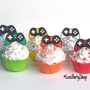 May include: Six cupcakes decorated with colorful frosting and black and white game controller toppers. The cupcakes are arranged in a row on a white surface. The cupcakes are decorated with sprinkles and edible glitter.