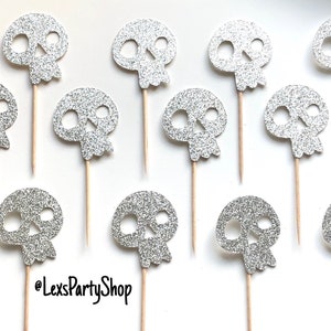 Może przedstawiać: Toppery na babeczki w kształcie czaszki z srebrnym brokatem na drewnianych patyczkach. Czaszki mają około 3,8 cm wysokości. @LexsPartyShop