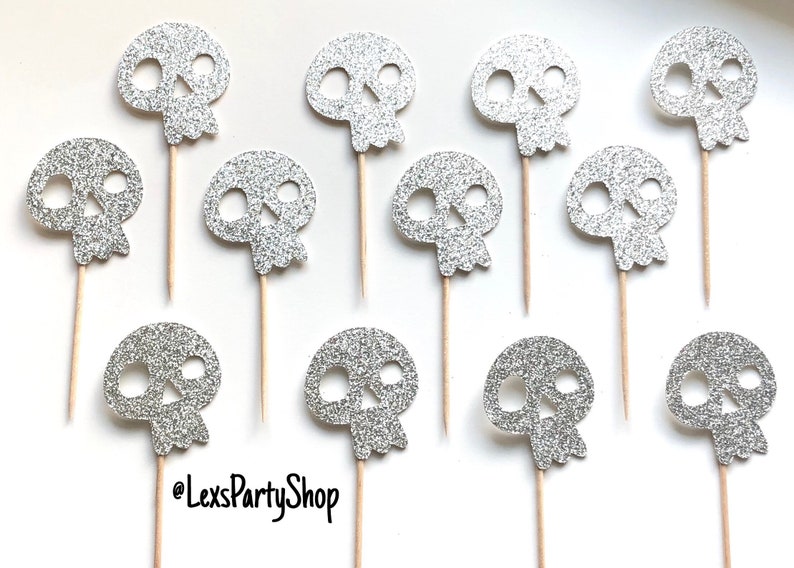Może przedstawiać: Toppery na babeczki w kształcie czaszki z srebrnym brokatem na drewnianych patyczkach. Czaszki mają około 3,8 cm wysokości. @LexsPartyShop