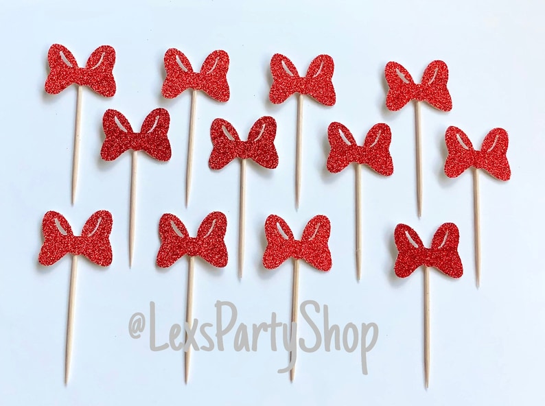 May include: Twelve red glitter bow cupcake toppers on wooden sticks. The bows are all the same size and shape, and they are arranged in a random pattern. The text "@LexsPartyShop" is visible in the bottom center of the image.