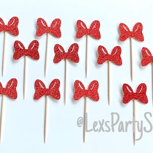 May include: Twelve red glitter bow cupcake toppers on wooden sticks. The bows are all the same size and shape. The background is white.