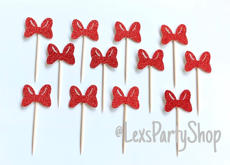 May include: Twelve red glitter bow cupcake toppers on wooden sticks. The bows are all the same size and shape. The background is white.
