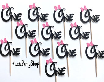 May include: Ten black glitter cupcake toppers with pink bows and the word "one" in a cursive font. The toppers are on a white background.
