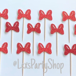 May include: Twelve red glitter bow cupcake toppers on wooden sticks. The bows are all the same size and shape, and they are arranged in a grid pattern. The background is white. @LexsPartyShop