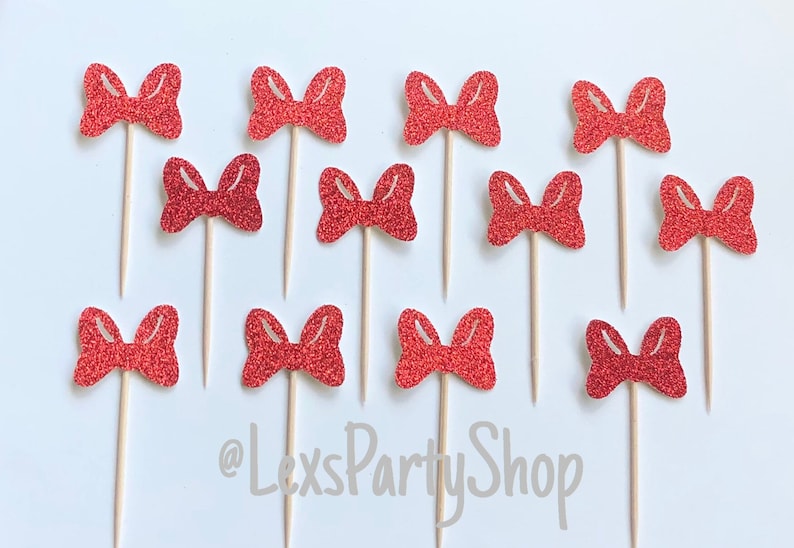 May include: Twelve red glitter bow cupcake toppers on wooden sticks. The bows are all the same size and shape, and they are arranged in a grid pattern. The background is white. @LexsPartyShop
