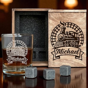 Puede incluir: Un vaso de whisky con un diseño de tren y el nombre "Michael" grabado, junto a una caja de madera con un diseño de tren a juego. Tres piedras de whisky grises están frente al vaso.