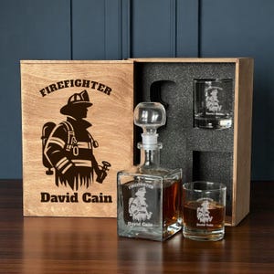 May include: A personalised firefighter gift set comprising a wooden box, a glass decanter, and two glasses. The decanter and glasses feature a firefighter graphic and the name "David Cain". The box is engraved with "Firefighter" and the same name.