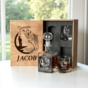 Könnte beinhalten: Ein hölzernes Geschenkbox-Set mit einer personalisierten Whisky-Karaffe und zwei Gläsern. Die Karaffe und die Gläser zeigen eine geätzte Angelszene mit dem Namen "JACOB". Die Box hat ein passendes graviertes Design.