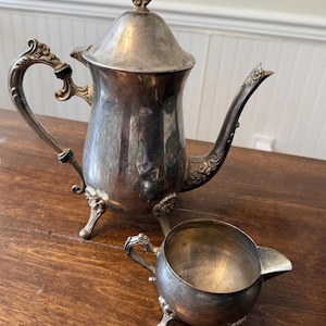 May include: A vintage silver tea set, comprising a teapot with ornate handle and lid detail, and a small creamer. The teapot features a curved spout and rests on three small feet. The creamer also has three feet and a small handle.