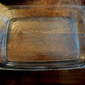 Puede incluir: Una fuente de horno rectangular de vidrio transparente con asas a cada lado. La fuente está vacía y se encuentra sobre una superficie de madera oscura. Las asas son rectangulares y están integradas en el diseño de la fuente.