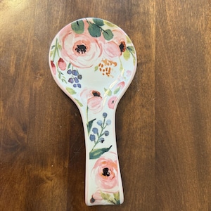 Puede incluir: Un reposacucharas de cerámica con un diseño floral. El reposacucharas es blanco con rosas rosas, hojas verdes y pequeñas bayas azules. Tiene una cabeza ancha y ovalada y un mango estrecho.