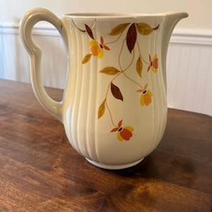 Puede incluir: Jarra de cerámica color crema con asa curva y boquilla. La jarra está decorada con un estampado floral de flores y hojas naranjas, amarillas y marrones. La jarra tiene detalles verticales acanalados.